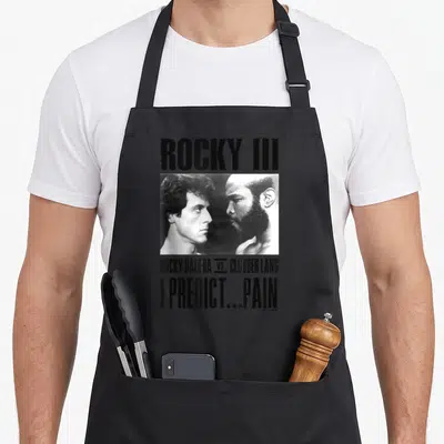 Tablier de cuisine noir avec une illustration monochrome de Rocky Balboa et Clubber Lang face à face, surplombée du texte Rocky III et I Predict... Pain.