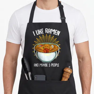 Tablier de cuisine noir avec illustration colorée d'un bol de ramen et texte blanc humoristique sur fond uni.