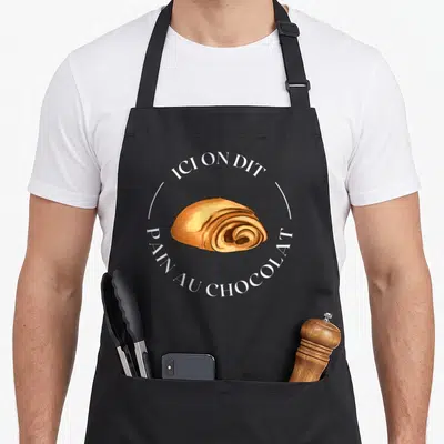 Tablier de cuisine noir avec illustration d'un pain au chocolat entouré du texte ici on dit pain au chocolat en blanc.