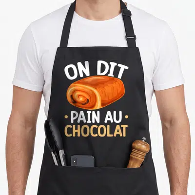 Tablier de cuisine noir avec illustration colorée d'un pain au chocolat et le texte On Dit Pain Au Chocolat imprimé en blanc et orange.