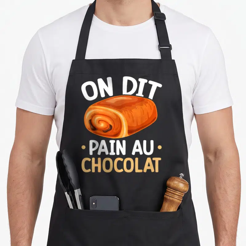 Tablier de cuisine noir avec illustration colorée d'un pain au chocolat et le texte On Dit Pain Au Chocolat imprimé en blanc et orange.