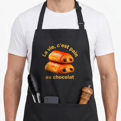 Tablier de cuisine noir avec deux pains au chocolat aux yeux et le texte La vie c'est pain au chocolat en jaune.