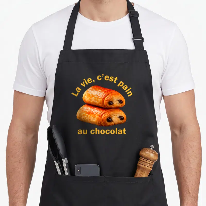 Tablier de cuisine noir avec deux pains au chocolat aux yeux et le texte La vie c'est pain au chocolat en jaune.