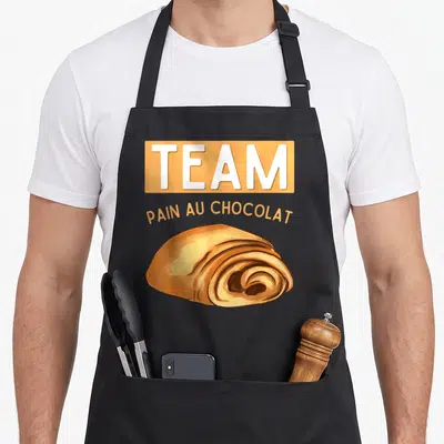 Tablier de cuisine noir avec l'inscription Team Pain au Chocolat et un dessin de viennoiserie dorée