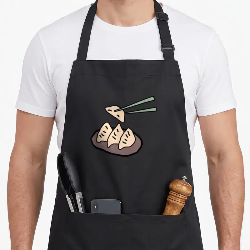 Tablier de cuisine noir avec illustration de gyoza sur assiette et baguettes vertes.