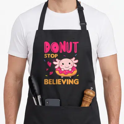 Tablier de cuisine noir avec un axolotl rose tenant un donut et le texte Donut Stop Believing en lettres colorées.