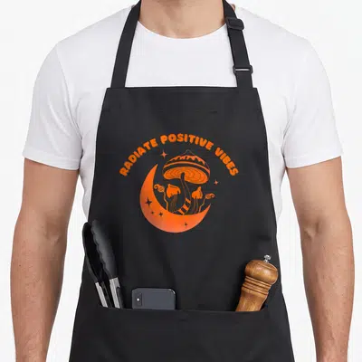 Tablier de cuisine noir avec illustration orange d'un champignon magique sous un croissant de lune et l'inscription Radiate Positive Energy.