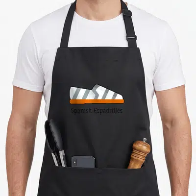 Tablier de cuisine noir avec illustration d'espadrille blanche et grise à base orange et texte Spanish Espadrilles en dessous.