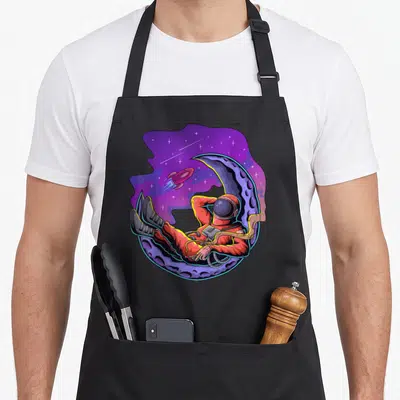 Tablier de cuisine noir avec illustration d'un astronaute reposant sur un croissant de lune sous un ciel galactique violet.