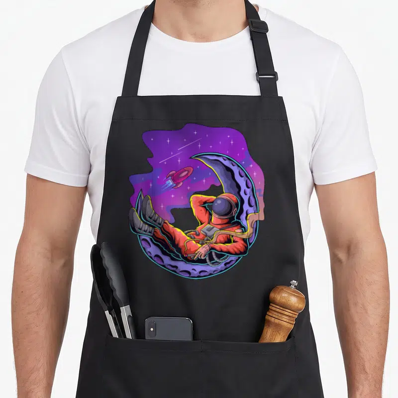Tablier de cuisine noir avec illustration d'un astronaute reposant sur un croissant de lune sous un ciel galactique violet.