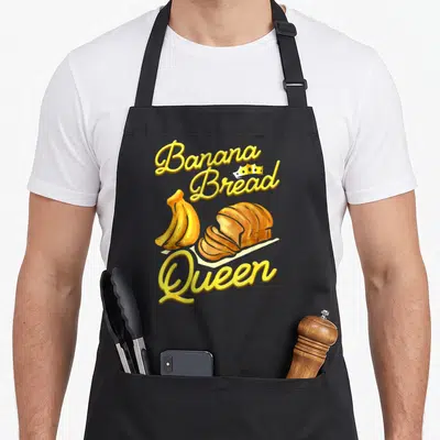 Tablier de cuisine noir avec illustration de bananes, pain tranché et texte Banana Bread Queen en jaune.