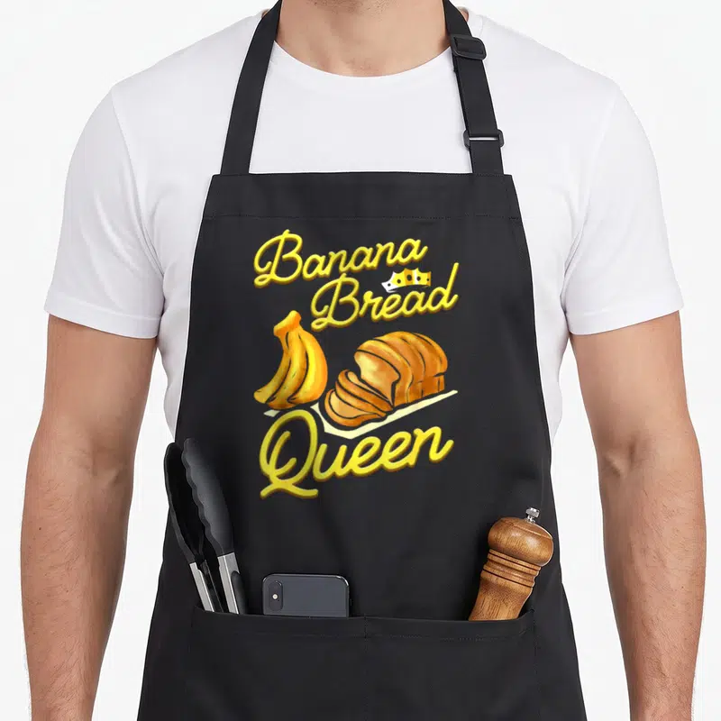 Tablier de cuisine noir avec illustration de bananes, pain tranché et texte Banana Bread Queen en jaune.