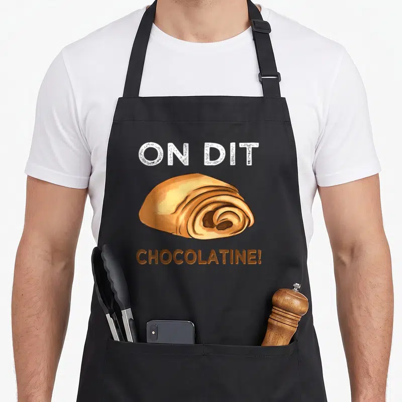 Tablier de cuisine noir avec une illustration de chocolatine et le texte On dit Chocolatine la boulangerie en lettres blanches et brunes.