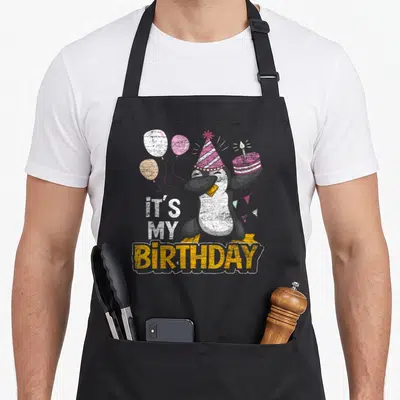 Tablier de cuisine noir avec illustration d'un pingouin en fête portant une casquette et tenant un gâteau ainsi que des ballons colorés.