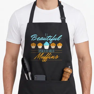 Tablier de cuisine noir avec cinq muffins dont un cupcake bleu et le texte Sois un beau petit gâteau.