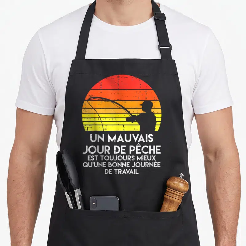 Tablier de cuisine noir avec silhouette de pêcheur et texte blanc sur fond de soleil rayé orange et jaune.