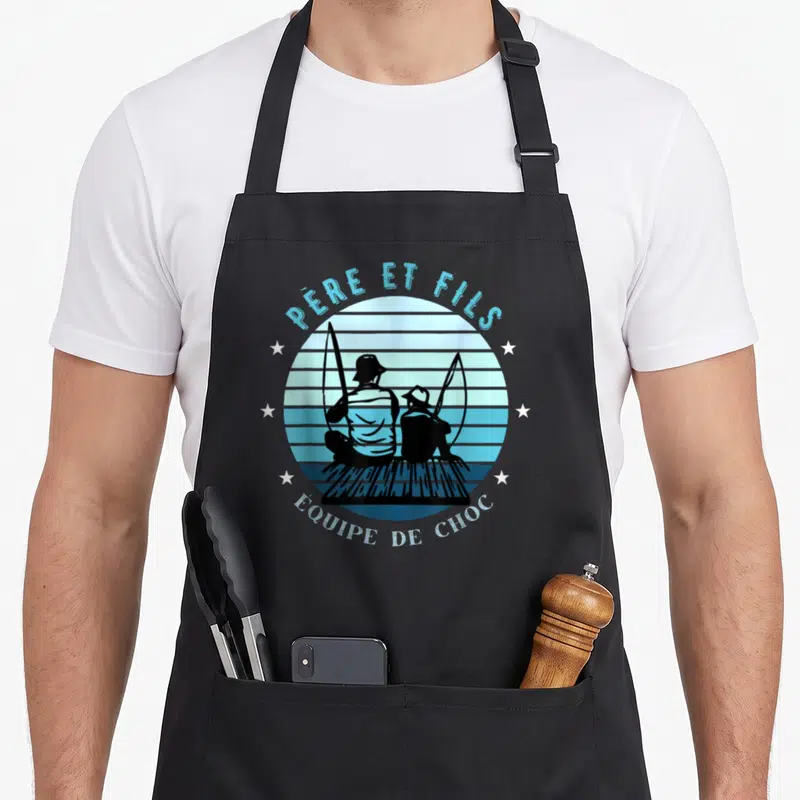 Tablier de cuisine noir avec une illustration circulaire représentant un père et un fils en silhouette sur fond de lignes bleues et de étoiles blanches.