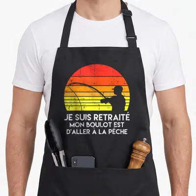 Tablier de cuisine noir avec une silhouette de pêcheur devant un soleil orange et le texte Je suis retraité mon boulot est d'aller à la pêche.