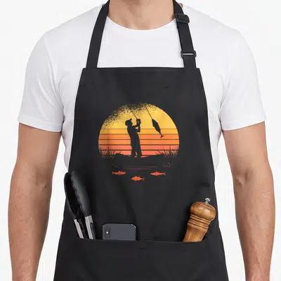 Tablier de cuisine noir avec silhouette d'un pêcheur tenant une canne à pêche et un poisson sautant sur fond de soleil couchant orange.