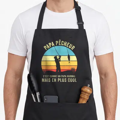 Tablier de cuisine noir avec silhouette de pêcheur et texte Papa Pêcheur mais en plus cool sur fond de soleil couchant.