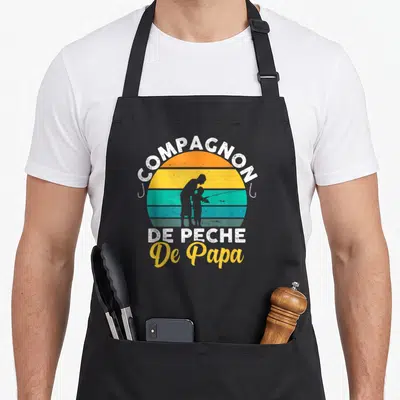 Tablier de cuisine noir avec illustration vintage d'un père et enfant pêchant sous un soleil multicolore et le texte Compagnon de pêche De Papa.