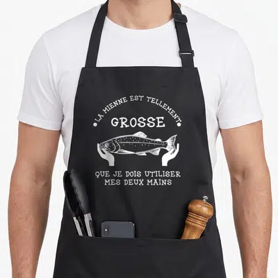 Tablier de cuisine noir avec illustration blanche d'un poisson tenu par deux mains et texte humoristique sur la taille du poisson.