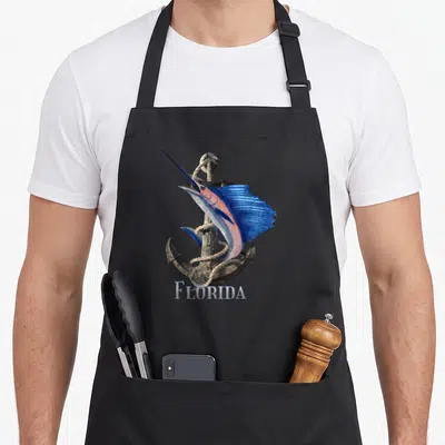 Tablier de cuisine noir avec un espadon bleu et rose enroulé autour d'une ancre métallique.