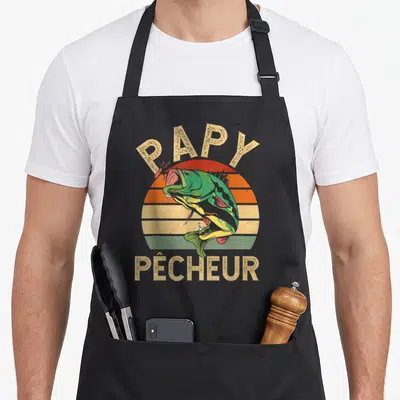 Tablier de cuisine noir avec inscription Papy Pêcheur et illustration colorée d'un poisson vert bondissant devant un soleil rayé.