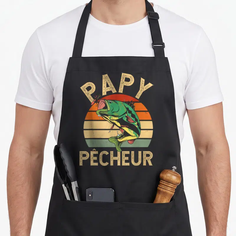 Tablier de cuisine noir avec inscription Papy Pêcheur et illustration colorée d'un poisson vert bondissant devant un soleil rayé.