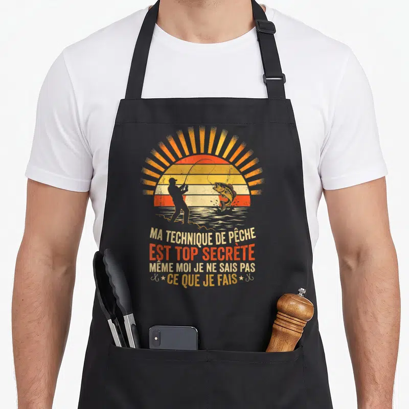Tablier de cuisine noir avec illustration d'un pecheur et d'un saumon devant un soleil couchant aux couleurs vintage orange et jaune, surmontant un texte humoristique en lettres capitales blanches et rouges.