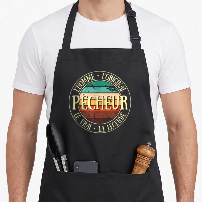 Tablier de cuisine noir avec logo circulaire rétro affichant Pêcheur LOriginal et La Légende sur fond de soleil coloré.