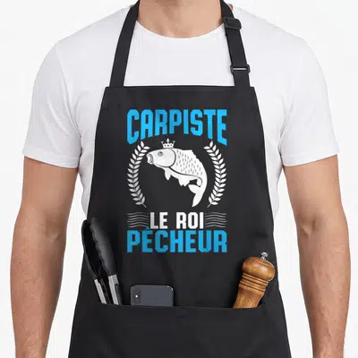 Tablier de cuisine noir avec une carpe blanche couronnée encadrée de lauriers et le texte Carpiste Le Roi Pêcheur en bleu et blanc.