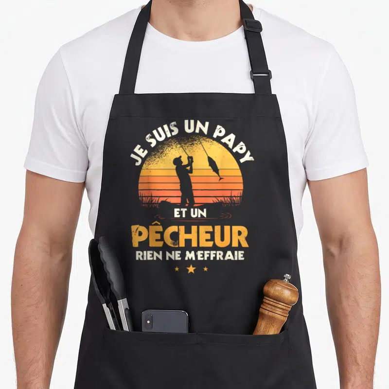 Tablier de cuisine noir avec silhouette de pêcheur et texte Je suis un papy et un pêcheur sur fond de soleil orange.