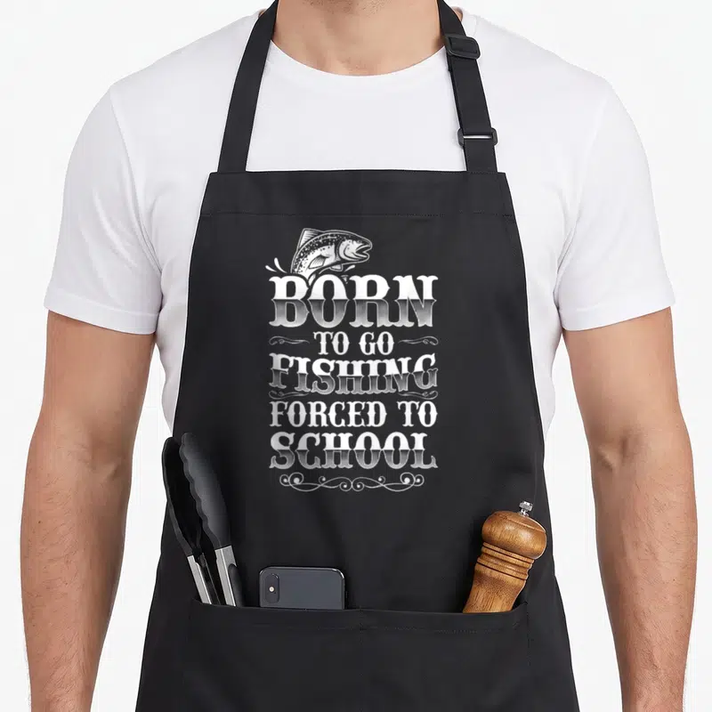 Tablier de cuisine noir avec illustration de poisson et texte Born To Go Fishing Forced To School en typographie blanche vintage.