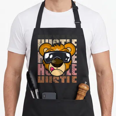 Tablier de cuisine noir avec tête de lion portant des lunettes noires et le texte HUSTLE en arrière-plan texturé.