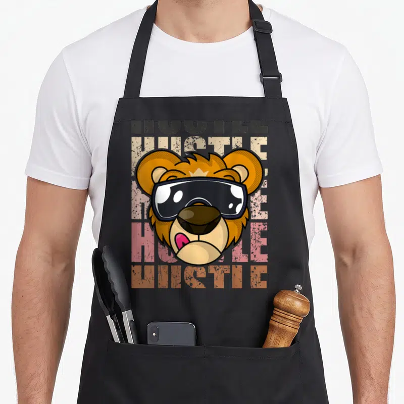 Tablier de cuisine noir avec tête de lion portant des lunettes noires et le texte HUSTLE en arrière-plan texturé.