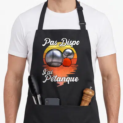 Tablier de cuisine noir avec inscription Pas Dispo J'ai Pétanque et illustration de deux boules métalliques devant un soleil orange.