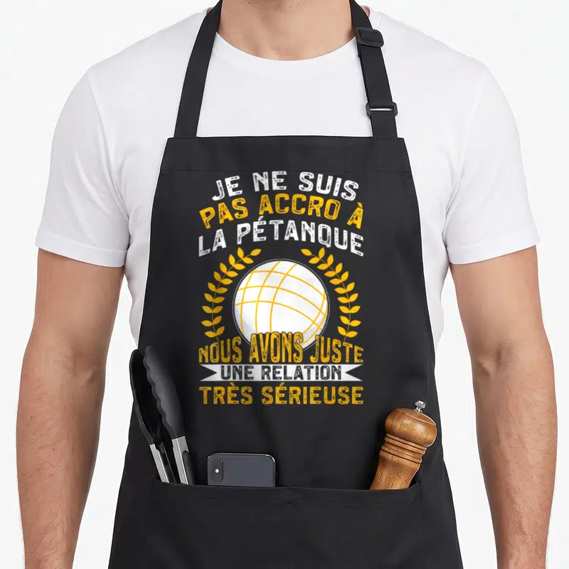 Tablier de cuisine noir avec une boule de pétanque encadrée de couronnes et un texte humoristique en blanc et orange.