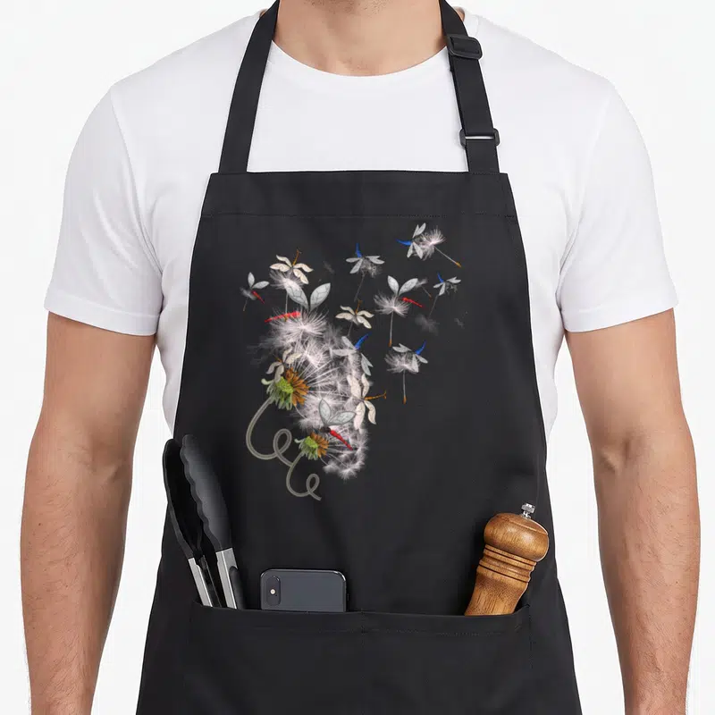 Tablier de cuisine noir avec des libellules aux ailes bleues et rouges s'envolant autour de tiges de pissenlit soufflées.