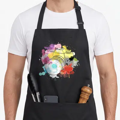 Tablier de cuisine noir avec illustration de caméra vintage entourée d'éclaboussures de peinture multicolore.