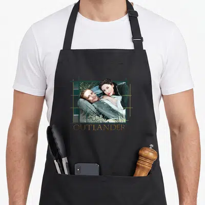 Tablier de cuisine noir avec illustration d'un couple allongé dans l'herbe encadré par des lignes vertes et le mot Outlander en doré.