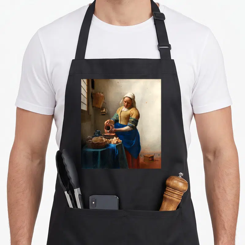 Tablier de cuisine noir avec reproduction de La Laitière de Vermeer imprimée sur le devant.