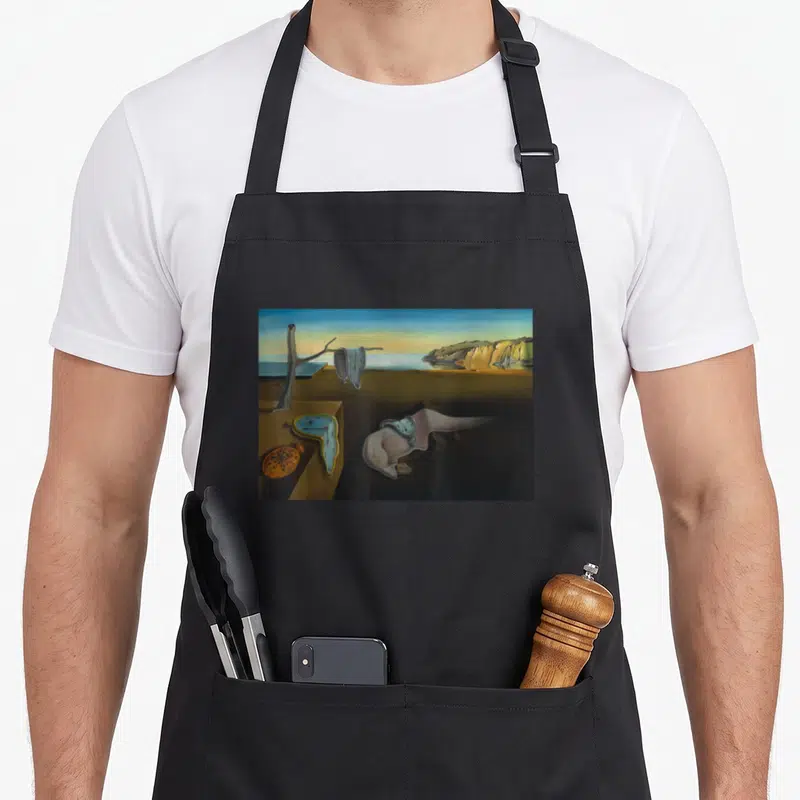 Tablier de cuisine noir avec reproduction de La persistance de la mémoire de Dali montrant des montres molles sur un paysage désertique.