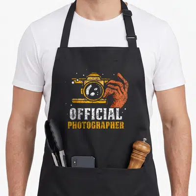 Tablier de cuisine noir avec illustration orange d'une main tenant un appareil photo et le texte official photographer en typographie usée.