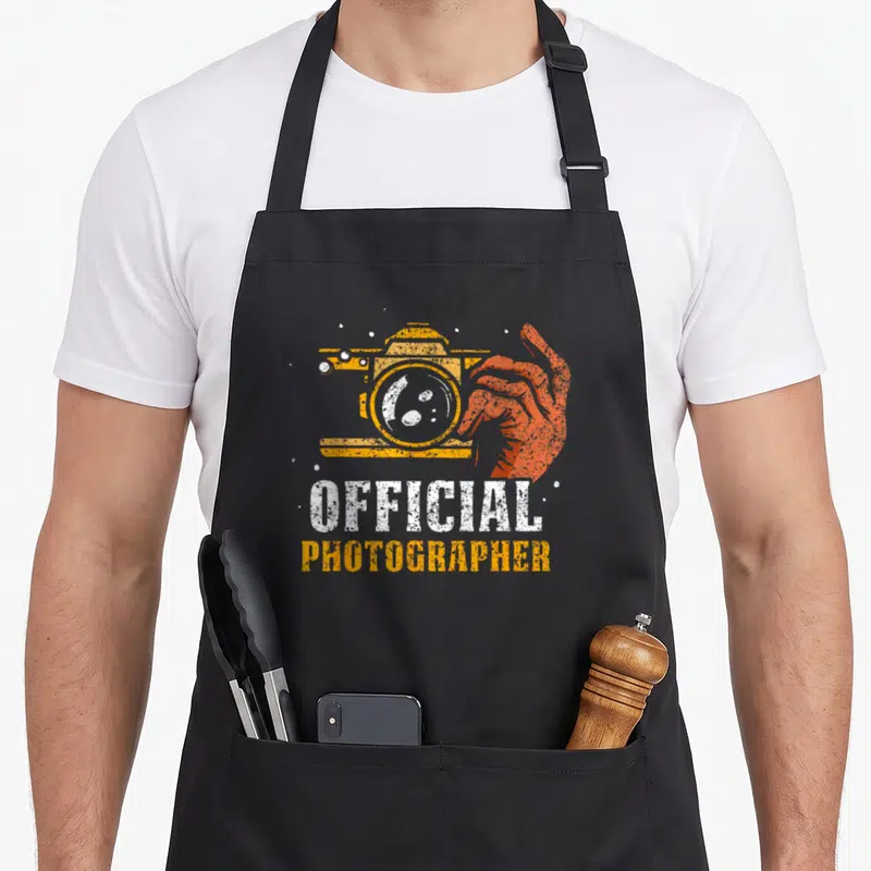 Tablier de cuisine noir avec illustration orange d'une main tenant un appareil photo et le texte official photographer en typographie usée.