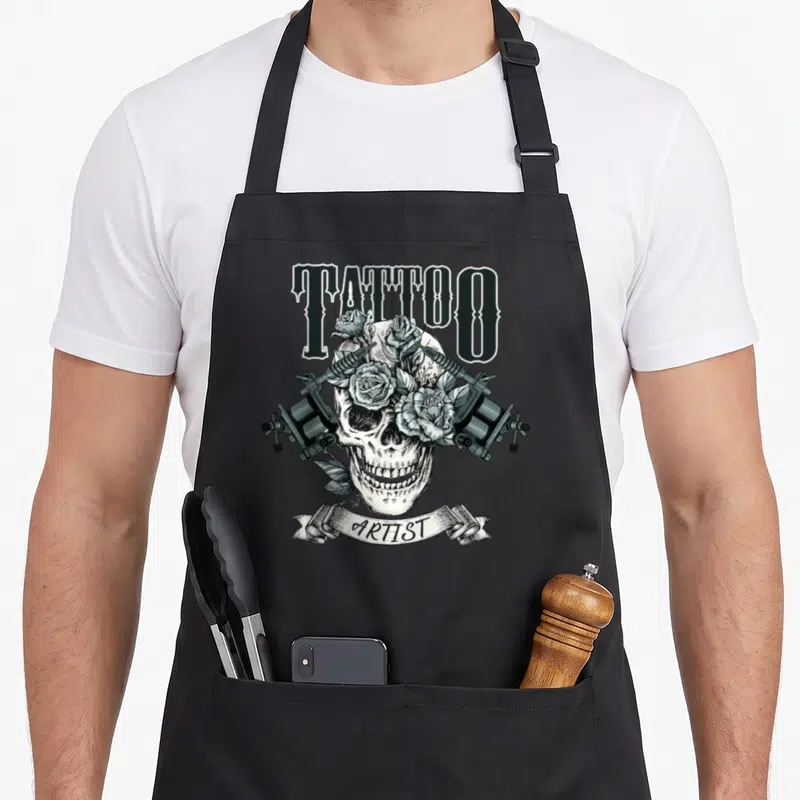 Tablier de cuisine noir avec un crâne, des roses et deux machines à tatouer croisées.
