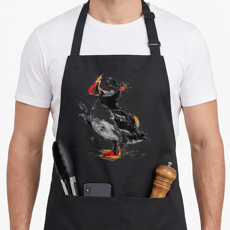 Tablier de cuisine noir avec un macareux dessiné au bec rouge et aux traits graphiques abstraits.