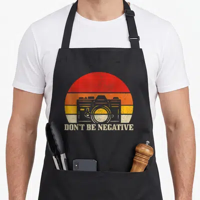 Tablier de cuisine noir avec illustration caméra vintage et texte Don't Be Negative sur fond de soleil rayé orange jaune beige