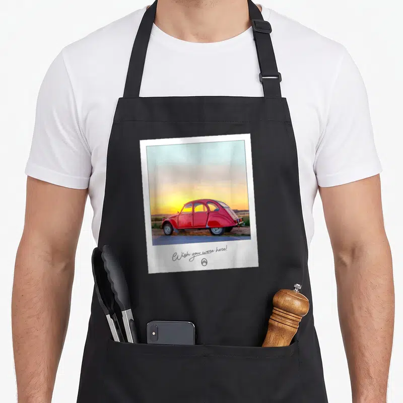 Tablier de cuisine noir avec une illustration polaroid d'une 2CV rouge au coucher du soleil et le texte Wish you were here en bas.