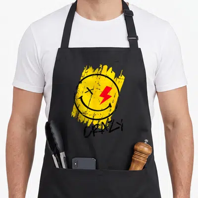 Tablier de cuisine noir avec un visage jaune sourant, un éclair rouge et le mot Crazy en bas.
